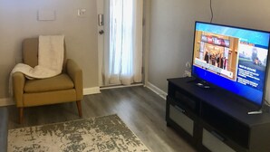 A living room with a beige armchair, a television displaying news, a patterned rug, and a door with curtains.