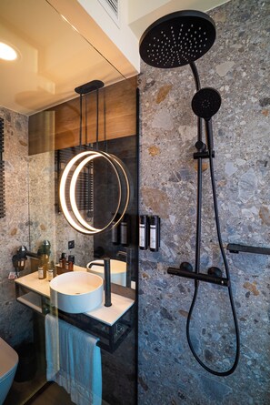 A modern bathroom with a stone wall, a round mirror with a light, a black showerhead, and a white sink. - Akeah Hotel Gran Via (Madrid)