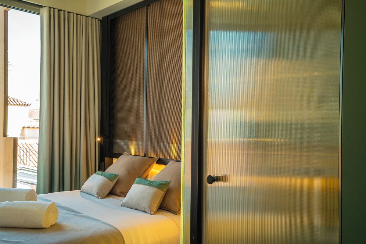 A hotel room with a large bed, a view through a window, and a door with a metal handle.