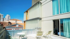 Un balcón con mesa y sillas, vista panorámica de la ciudad y un edificio con amplias ventanas.