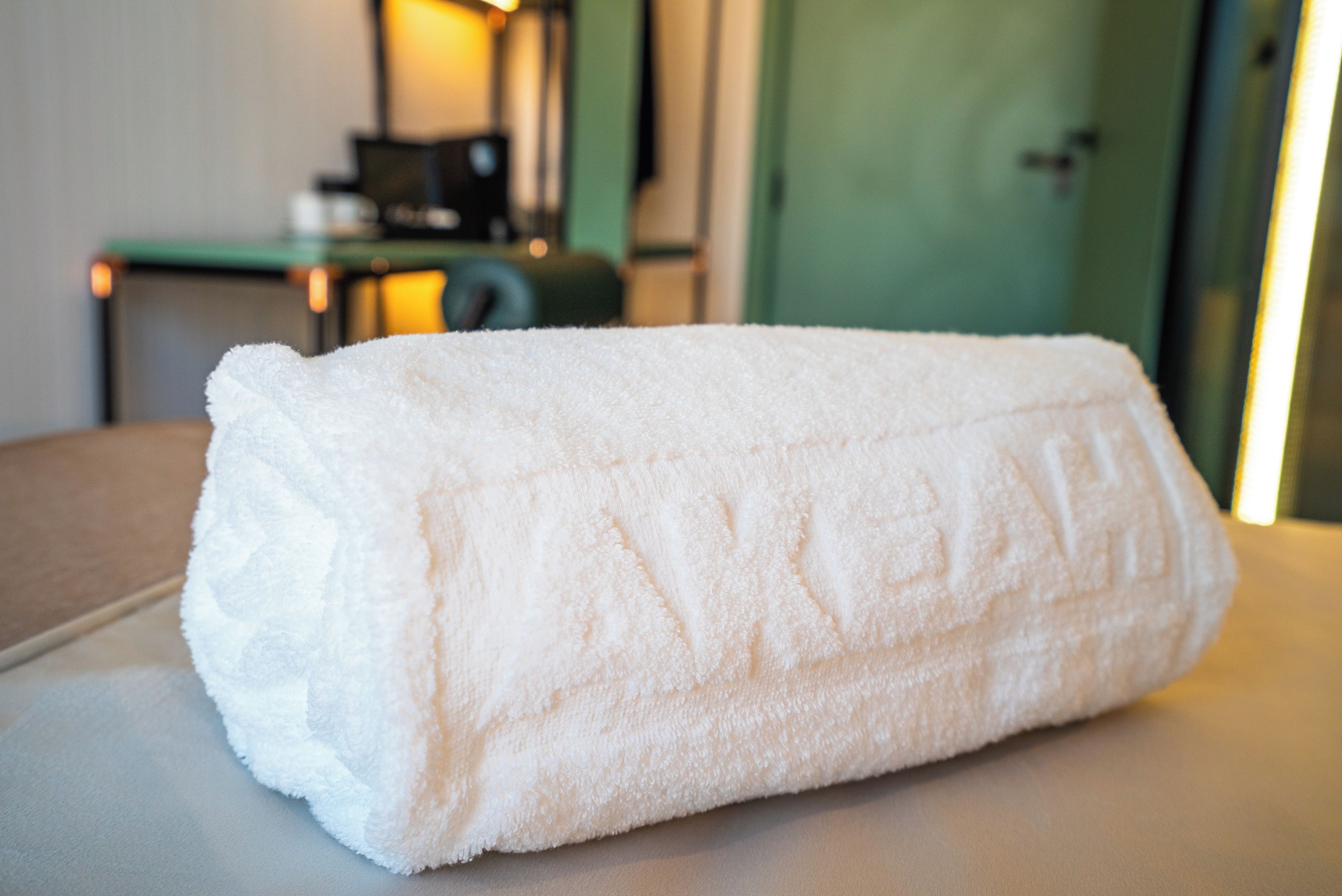 A neatly folded white towel on a table in a room with a green door and a small table with a lamp in the background.