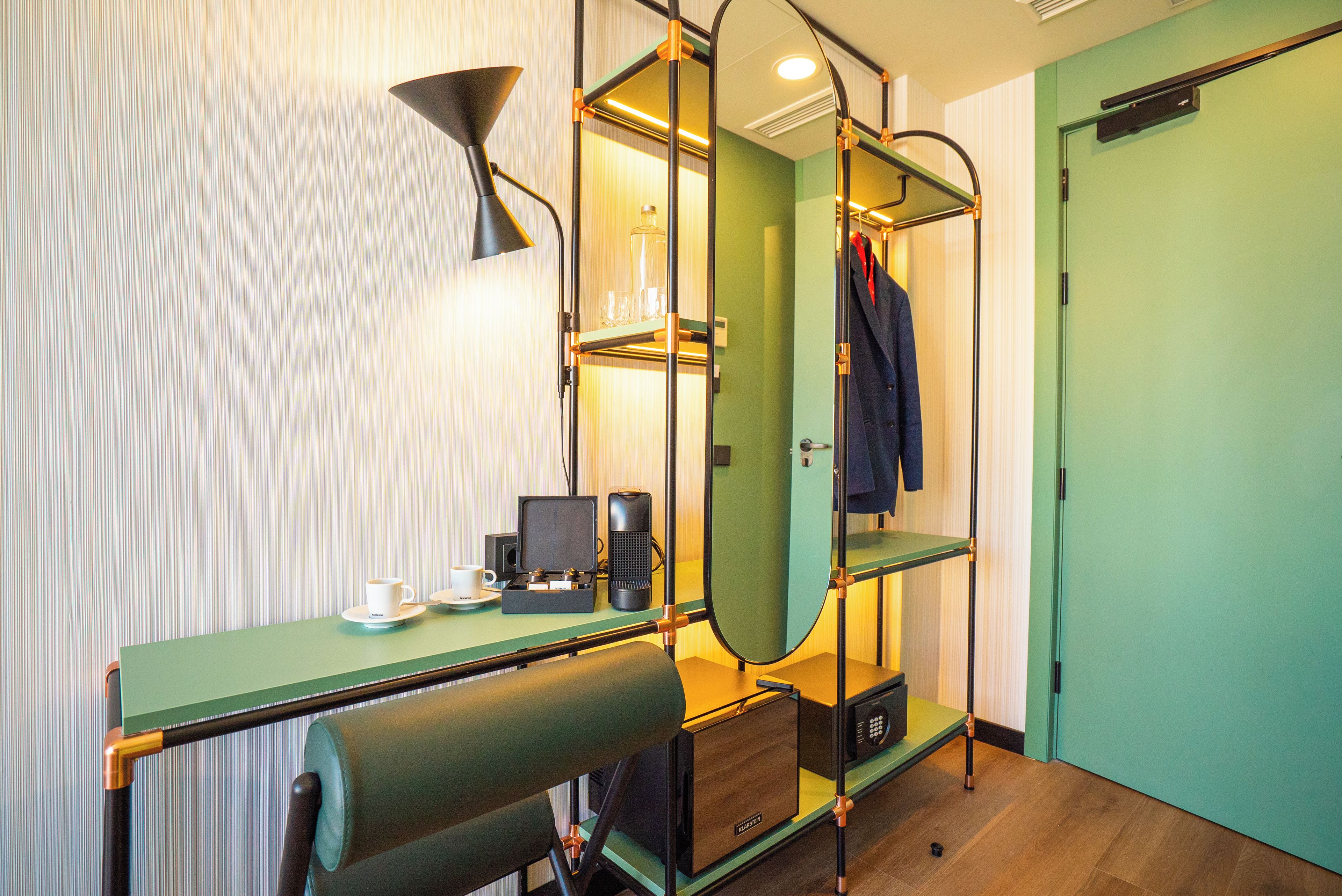 A compact hotel room with a green desk, a mirror, a coat rack, and a green door.
