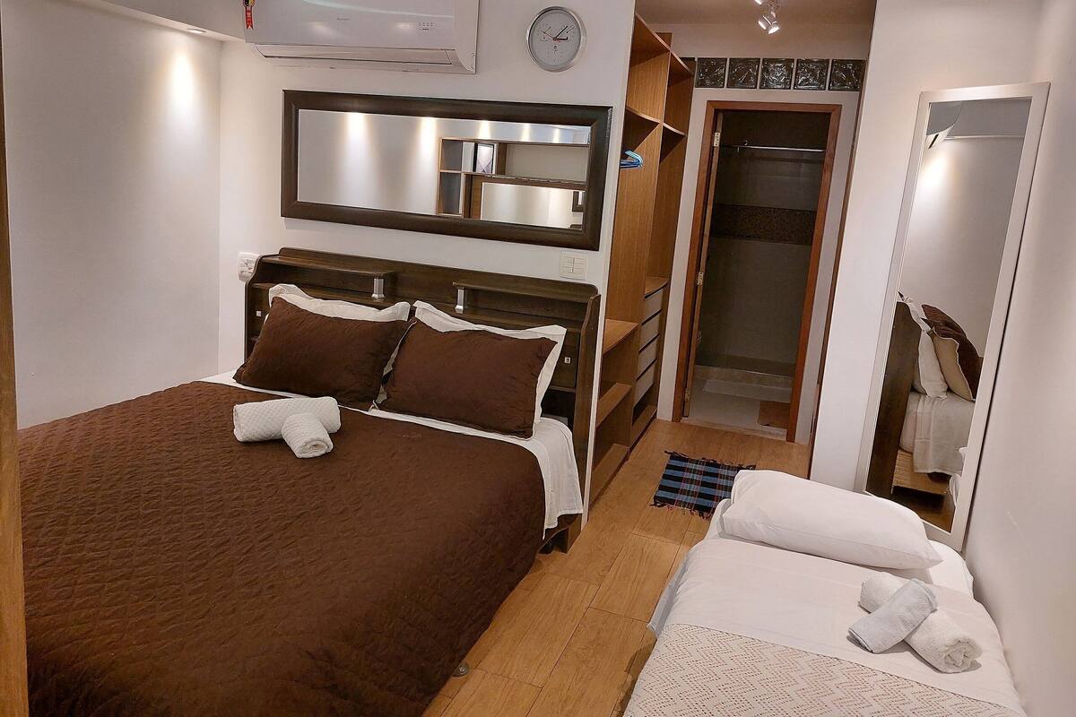 A bedroom with a large bed, a bedside table, a mirror, a wardrobe, and a shower area.