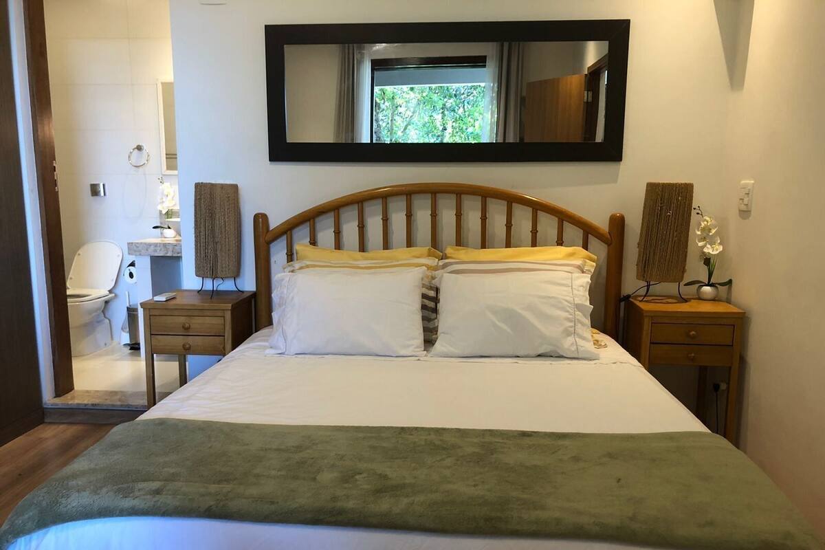 A bedroom with a bed, a wooden headboard, a bedside table, a nightstand, a mirror, and a bathroom visible through an open door.
