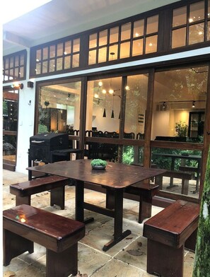 An outdoor patio area with a wooden table and benches, large windows, and a grill.