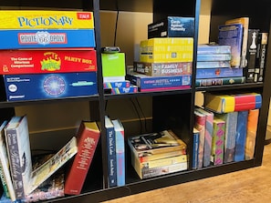 A bookshelf with various books and games, including 'Pictionary', 'Route 66', 'Apples to Apples', 'Party Box', 'Millionaire', 'Uttter Nonsense!', 'Cards Against Humanity', 'Vintage Motel Sogos', and several novels. - Blue Mountain Cabin Escape/AC/Pet Friendly (Flagstaff)