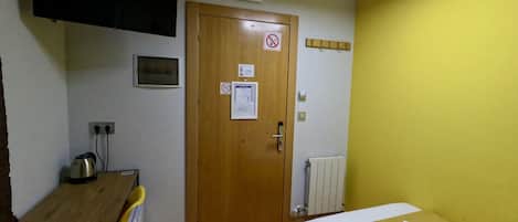 A small room with a wooden desk, a bed with a white blanket, a yellow wall, a door with a no smoking sign, and a ceiling light.