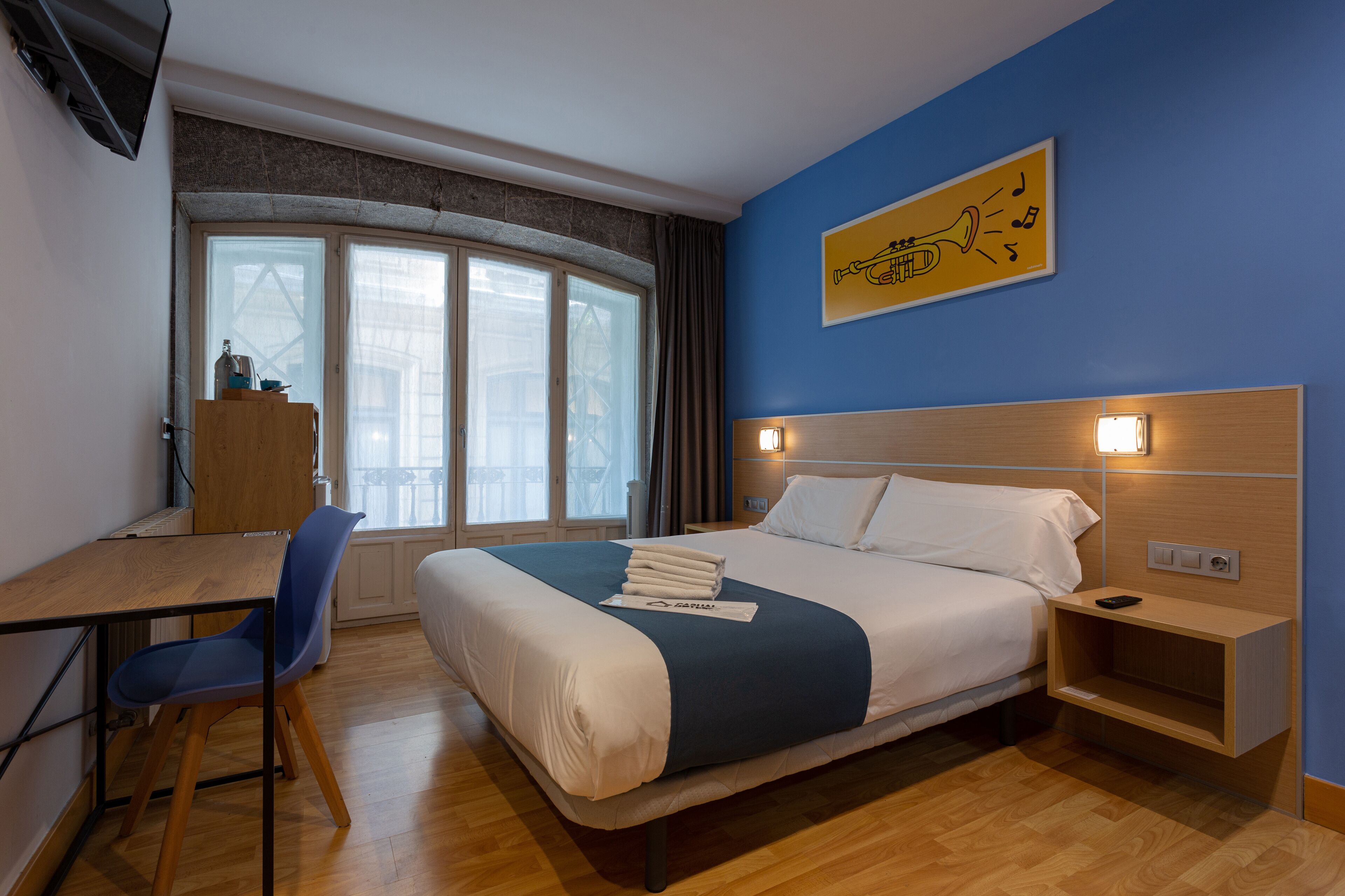 A hotel room with a bed, a desk, a chair, a nightstand, a wall-mounted TV, and a window with curtains.
