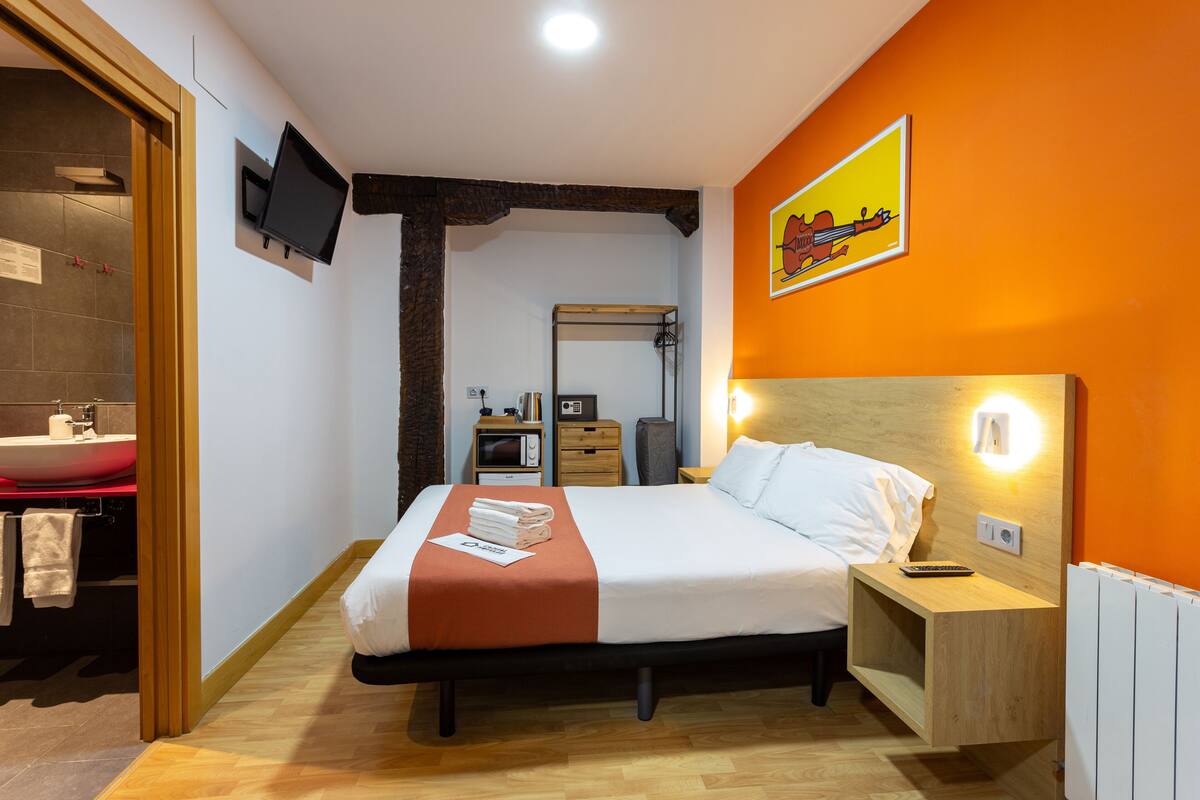 Una habitación de hotel con cama, mesita de noche, televisión y baño con lavabo y espejo.