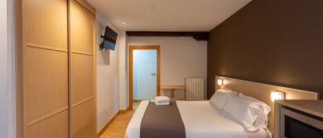A hotel room with a bed, bedside TV, wooden wardrobe, and a door leading to another room.