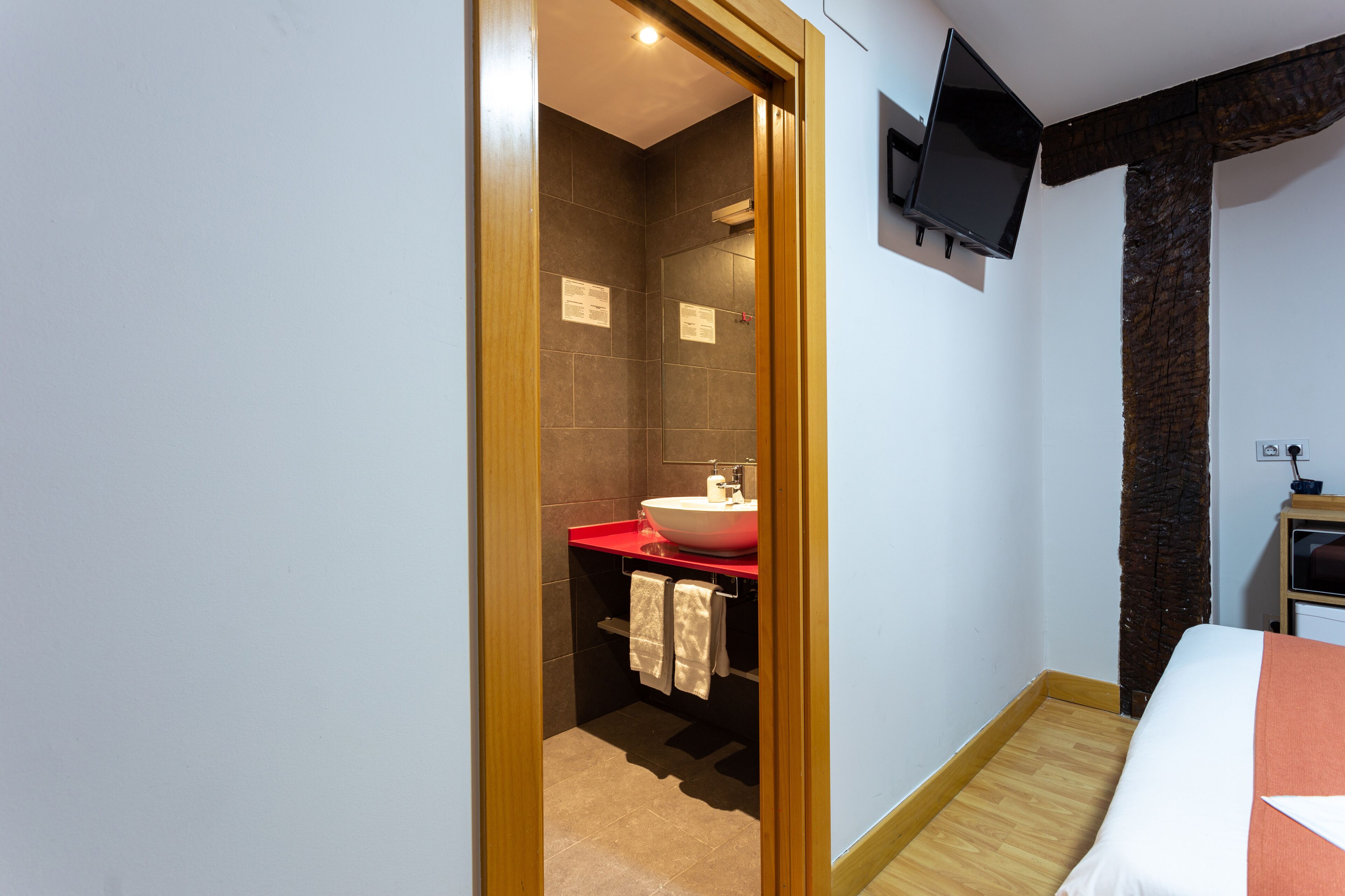 Une chambre d’hôtel avec un plancher en bois, un lit avec une literie blanche et orange, un lavabo rouge et un téléviseur fixé au mur.