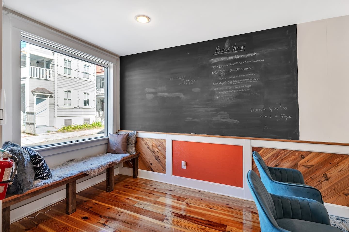 A room with a chalkboard displaying a schedule, a bench, a chair, and a window with a view of buildings.