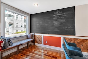 Quarto com quadro negro exibindo um cronograma, um banco, uma cadeira e uma janela com vista para edifícios.