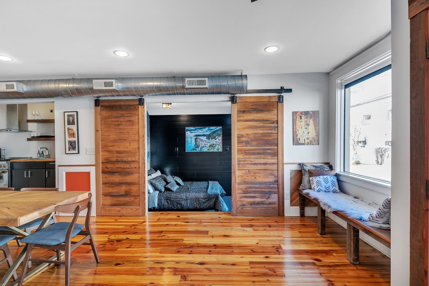 A modern interior with a wooden floor, a dining area, a living space with a sofa and a bench, and a kitchen in the background.