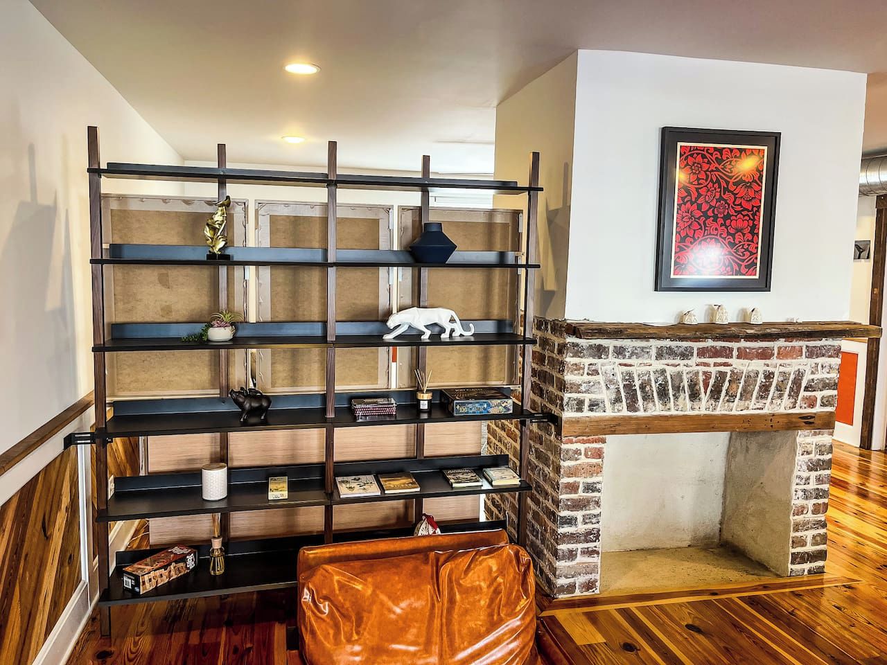 A room with a brick fireplace, a black metal shelving unit with various items, and a brown leather ottoman.