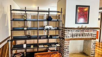 A room with a brick fireplace, a black metal shelving unit with various items, and a brown leather ottoman.