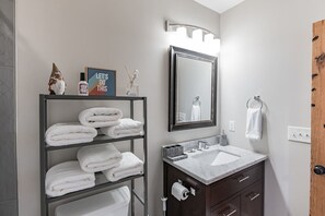 A modern bathroom with a vanity, a shelf with folded towels, a mirror with lights, and a wooden door. - Timeless 2 BR Home | Heart of Charleston | King St (Charleston)