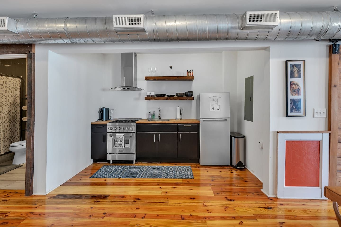 Cocina compacta con estufa, horno y refrigerador, piso de madera y conductos de ventilación visibles.