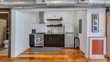 Cocina compacta con estufa, horno y refrigerador, piso de madera y conductos de ventilación visibles.