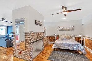 A modern bedroom with a brick fireplace, a bed with patterned bedding, a ceiling fan, and a view of the living area. - Timeless 2 BR Home | Heart of Charleston | King St (Charleston)