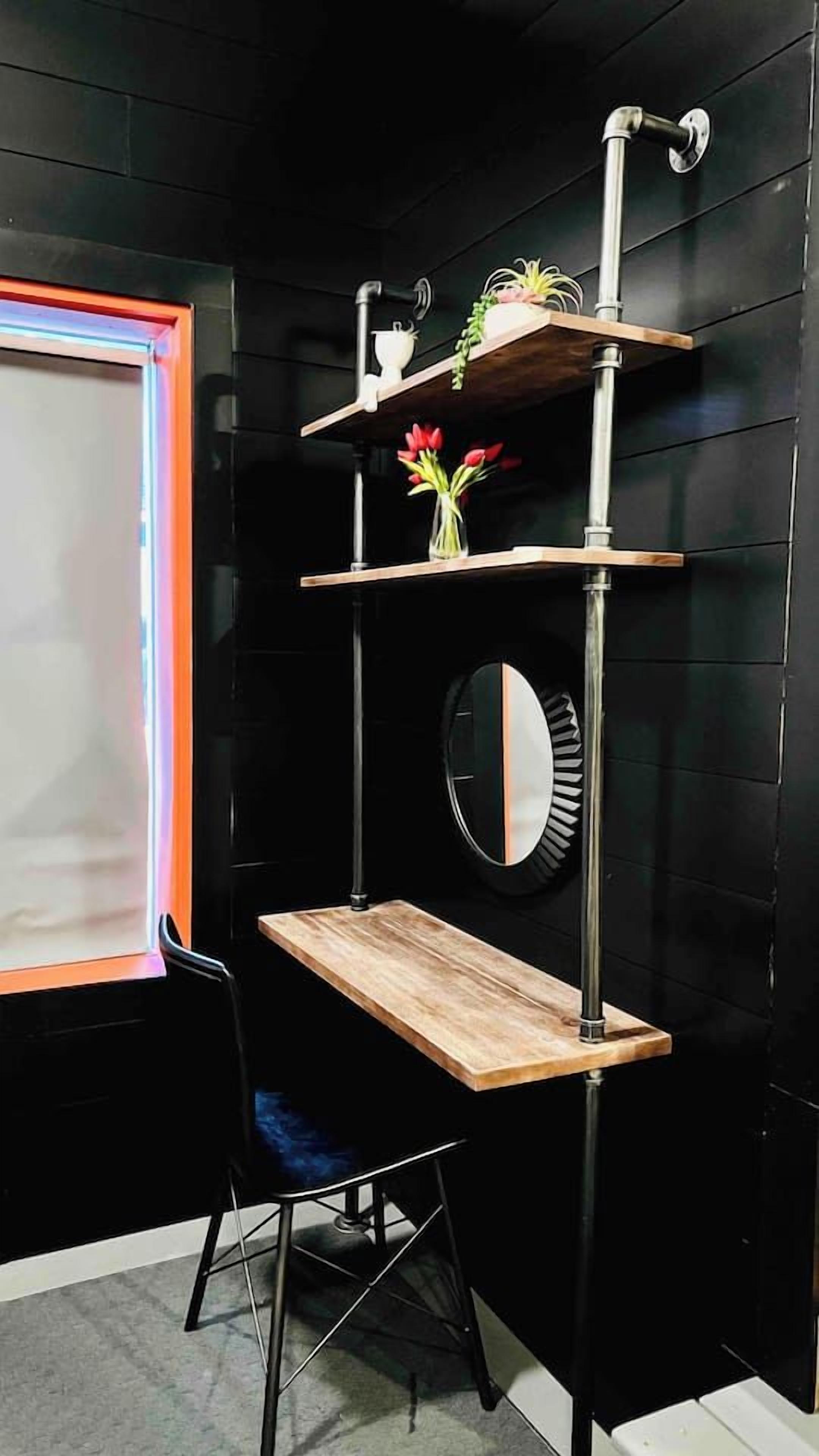 Una habitación con pared negra, un escritorio de madera, una silla, una estantería con plantas y un espejo.