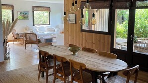 A dining area with a wooden table and chairs, a hanging light fixture, and a view of the outdoors through a large window. - WOODEN HOUSE TYPICAL OF THE BASIN (Lège-Cap-Ferret)