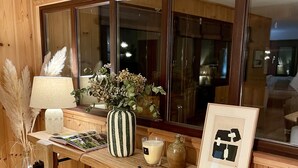 A wooden room with a large window, a wooden bench, a vase with dried flowers, a candle, and a framed picture. - WOODEN HOUSE TYPICAL OF THE BASIN (Lège-Cap-Ferret)