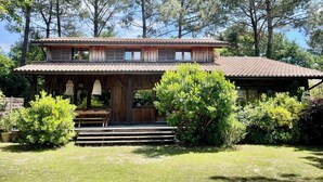 A rustic two-story wooden house with a tiled roof, surrounded by lush greenery and tall trees. - WOODEN HOUSE TYPICAL OF THE BASIN (Lège-Cap-Ferret)