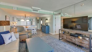 Ein Wohnzimmer mit Fernseher, Sofa und Couchtisch, ein Essbereich mit Stühlen und eine Küche im Hintergrund.