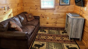 Une chambre confortable dans un chalet, avec un canapé en cuir, un petit meuble, une télévision et une fenêtre équipée de stores.