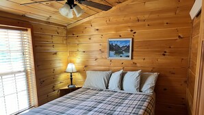 Une chambre dans un chalet en bois, avec un lit, une table de chevet, une lampe et une fenêtre avec des stores.