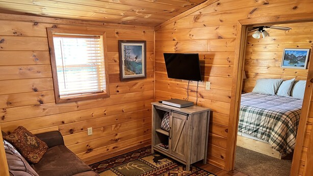 L’intérieur chaleureux d’un chalet avec un plancher en bois, un canapé en cuir marron, un téléviseur fixé au mur et un lit recouvert d’une couverture à carreaux.