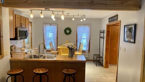 A kitchen with wooden cabinets, a stainless steel sink, and a bar area with stools. There are two windows with curtains, a wreath on the wall, and a sign that reads "DO NOT FEED THE BEARS".