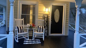 A porch with two white chairs, a small table with a vase of flowers, and a black door with a 'WELCOME' sign.