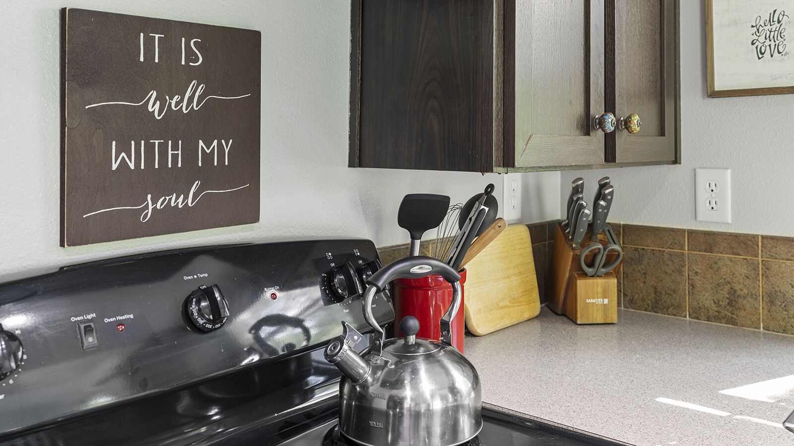 Una cocina con estufa, hervidor, una taza roja y un bloque de cuchillos. Un cartel en la pared dice "IT IS well with my soul".