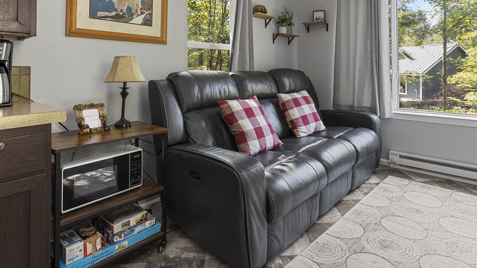 Un sofá de cuero negro con dos almohadas a cuadros rojos y blancos, una mesita con una lámpara, un microondas y una estantería con libros y objetos decorativos.