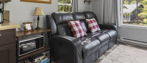 Un sofá de cuero negro con dos almohadas a cuadros rojos y blancos, una mesita con una lámpara, un microondas y una estantería con libros y objetos decorativos.