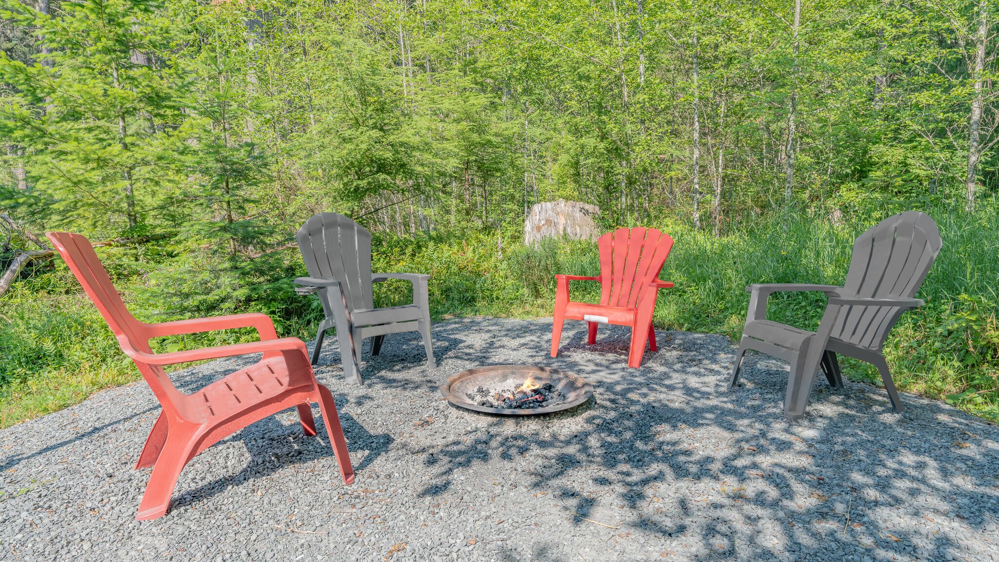 A fire pit surrounded by four Adirondack chairs in a forested area.