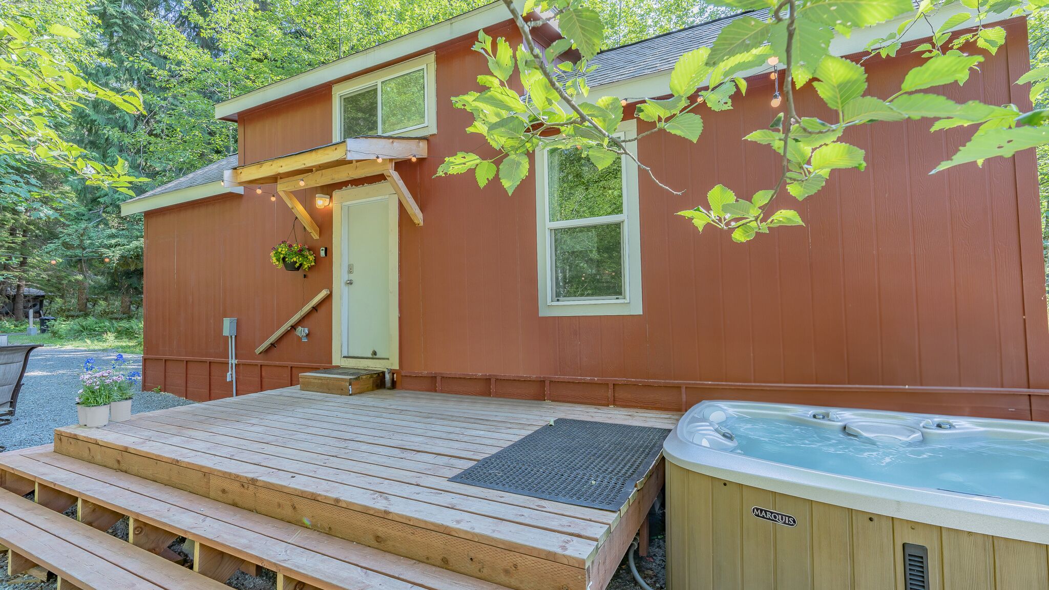 A wooden deck leading to a house with a hot tub, a small porch, and a window with a view of the surrounding trees.