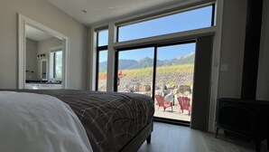 A bedroom with a large bed, a dresser, a mirror, and a sliding glass door leading to an outdoor area with chairs and a view of mountains. - Ski In to your Hot Tub & Sauna (Fernie)