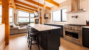 A modern kitchen with a central island, wooden beams, large windows, and a built-in stove. - Ski In to your Hot Tub & Sauna (Fernie)