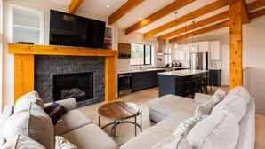 A modern living room with a fireplace, a television, and a kitchen area with a bar. - Ski In to your Hot Tub & Sauna (Fernie)