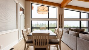 A spacious dining area with a large table and chairs, a sofa, and large windows offering a view of the outdoors. - Ski In to your Hot Tub & Sauna (Fernie)