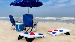 Strandaufbau mit blauem Sonnenschirm, zwei Liegestühlen, einer Kühlbox und einem kleinen weißen Tisch mit roten und blauen Kissen.