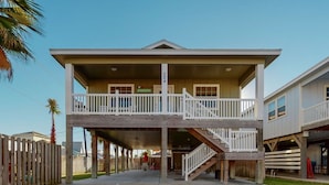 Ein zweistöckiges Strandhaus mit Balkon, überdachter Veranda und einer Treppe, die zum oberen Stockwerk führt.