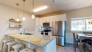 Una cocina con armarios blancos, un refrigerador de acero inoxidable, un microondas y una encimera de granito con individuales tejidos. Hay taburetes de bar y una ventana con persianas.