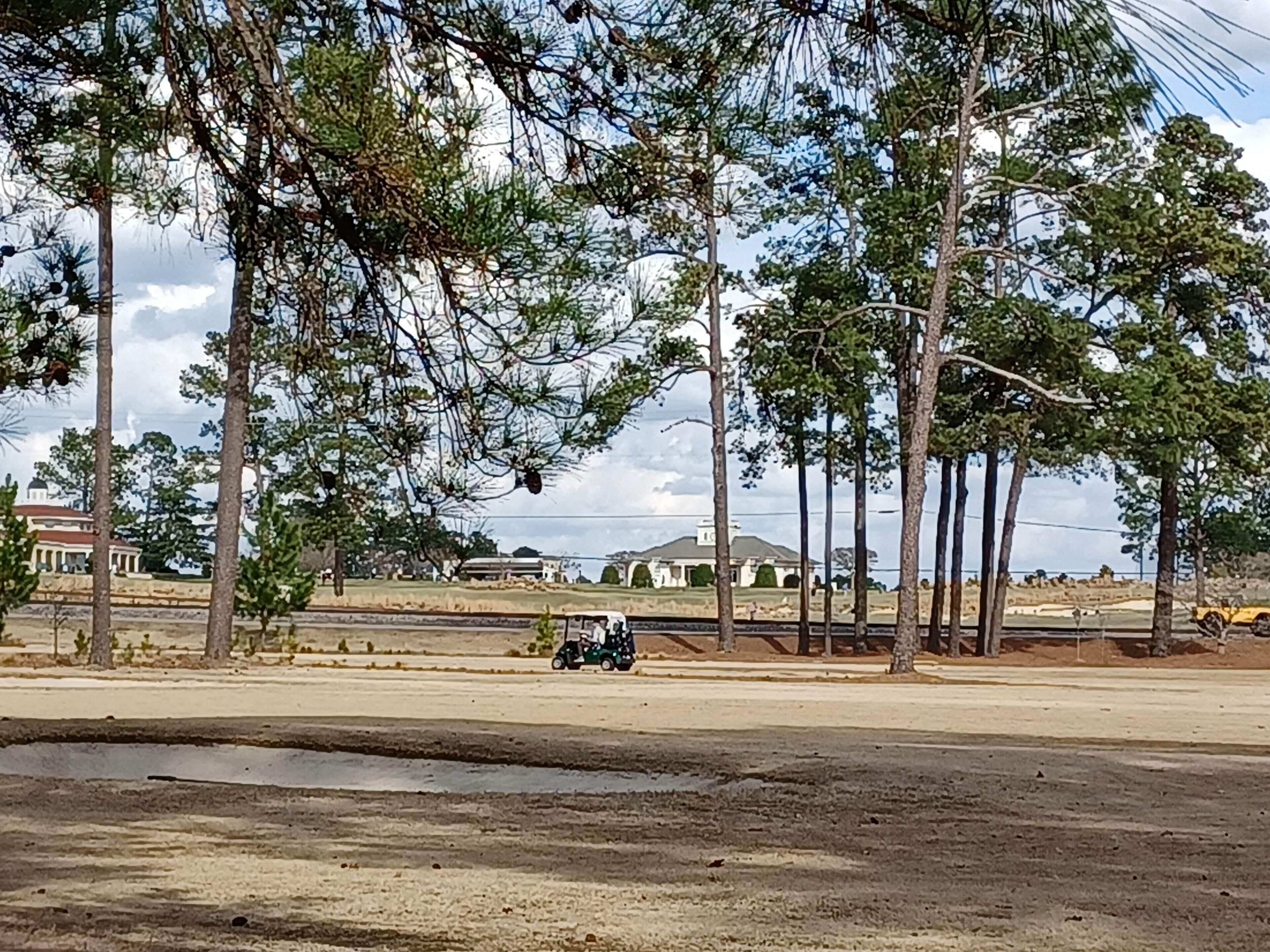 En parkeringsplats med några få fordon, omgiven av höga träd och en byggnad i bakgrunden.