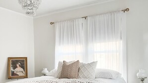A bedroom with a wooden bed, white bedding, a chandelier, a nightstand with a lamp, and a window with sheer curtains. - The Chandelier House of Midtown - Near It All! (Memphis)