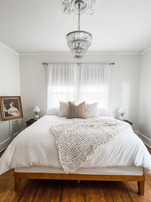 Een slaapkamer met een houten bed, witte beddengoed, een witte gebreide plaid, een kroonluchter en een lijst met een afbeelding aan de muur.