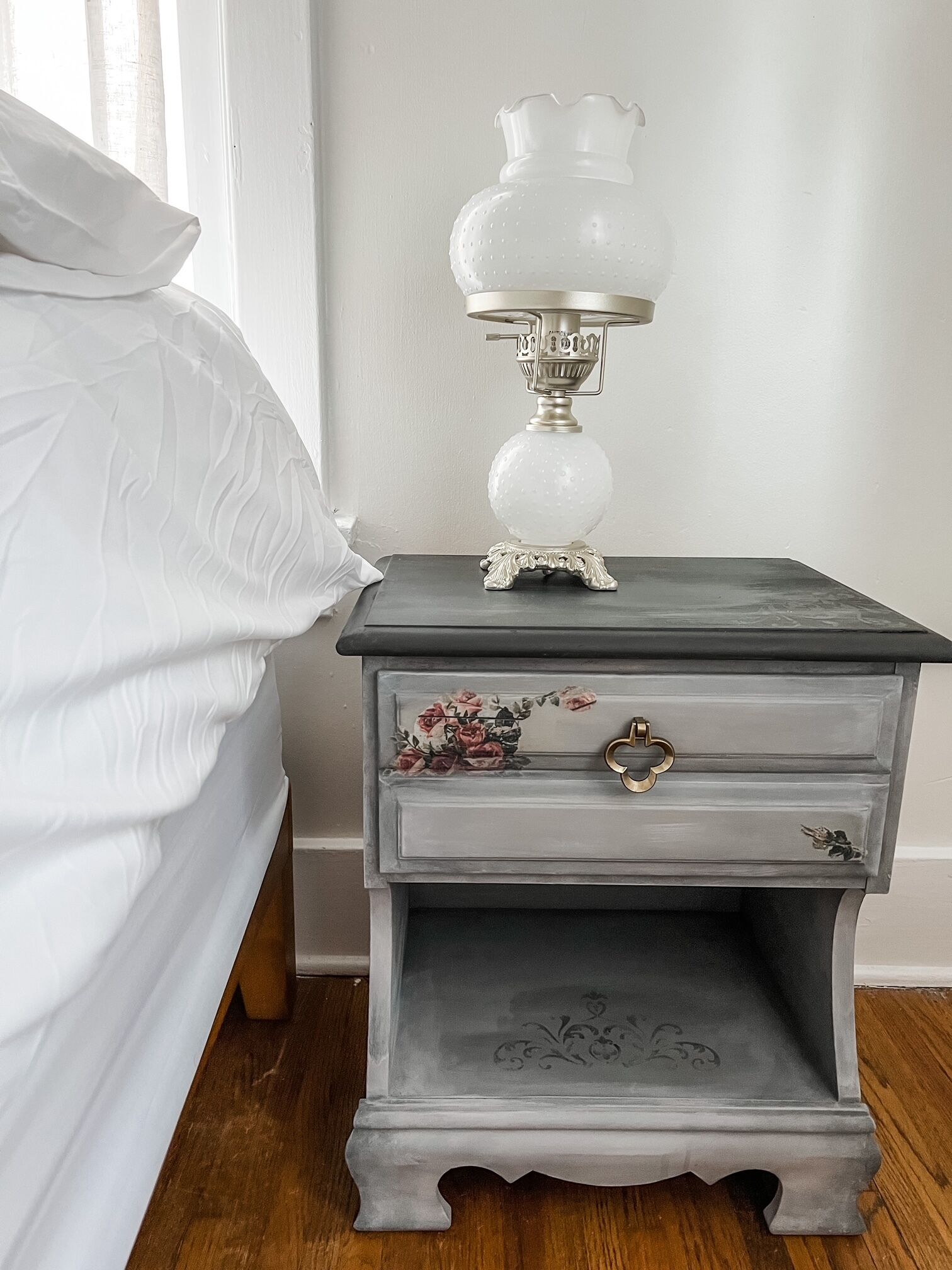Ein weißes Bett mit einem weißen Kopfkissen, ein grauer Nachttisch mit Blumenschublade und eine klare Glaslampe mit Metallfuß.